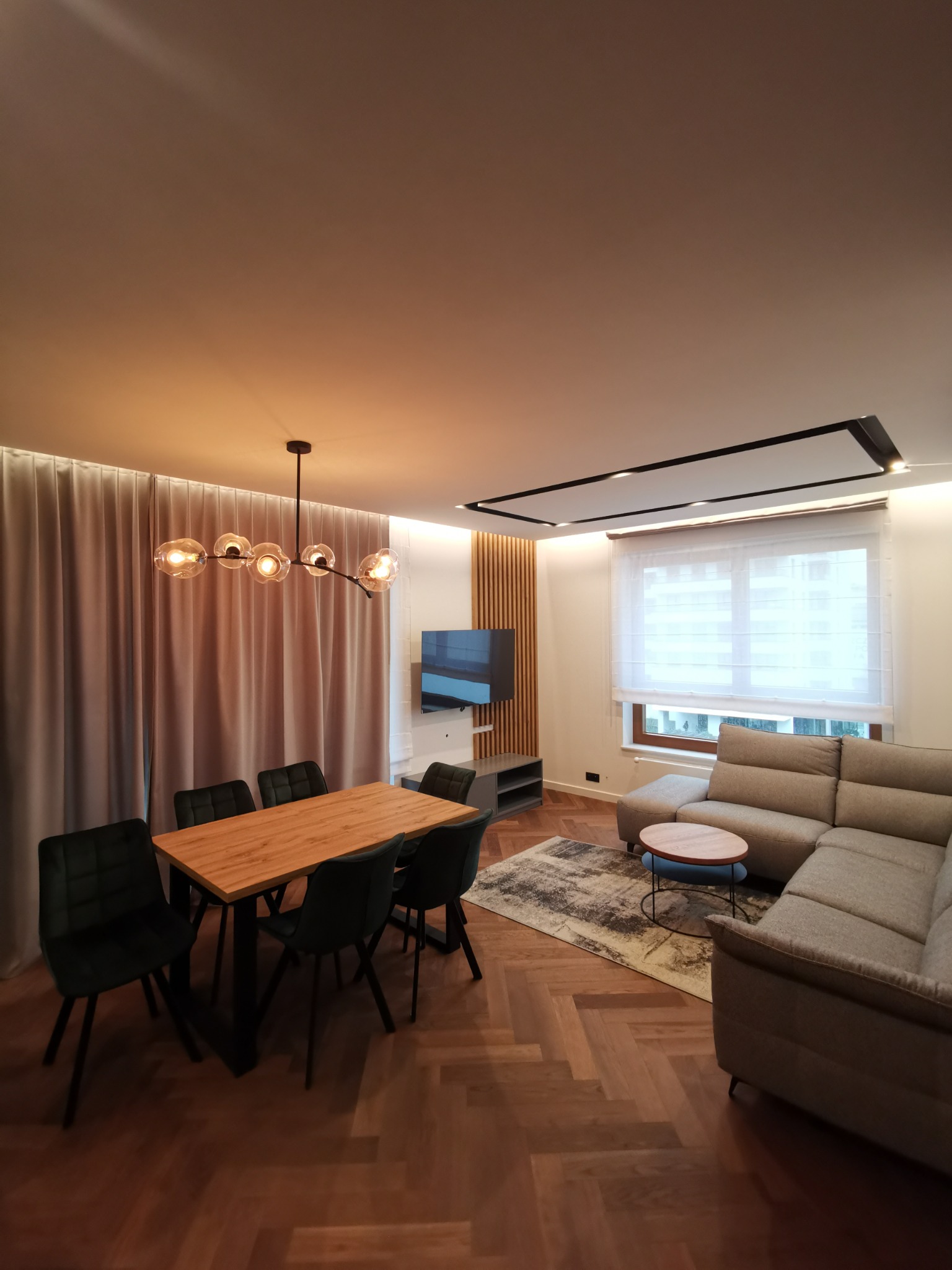 Wykończone wnętrze salonu z parkietem w jodełkę, szarą sofą narożną, drewnianym stołem z krzesłami i nowoczesną lampą wiszącą z szklanymi kloszami.