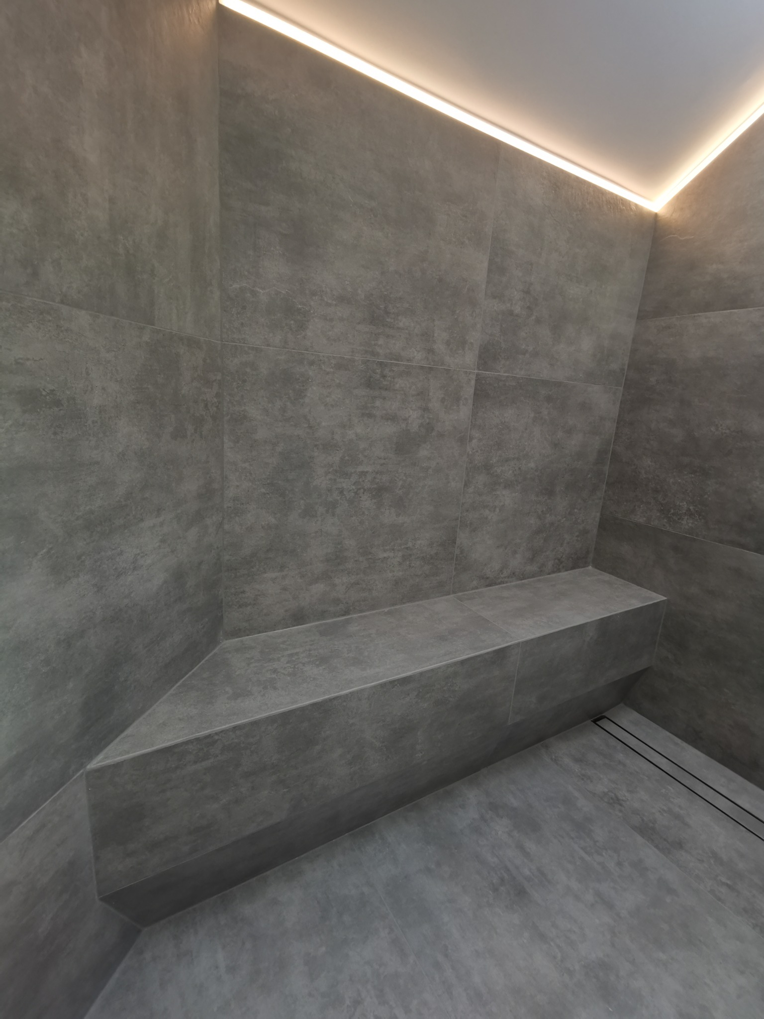 Nowoczesna kabina prysznicowa wyłożona szarymi płytkami imitującymi beton, z wbudowanym siedziskiem i liniowym odpływem, oświetlona subtelnym światłem LED.