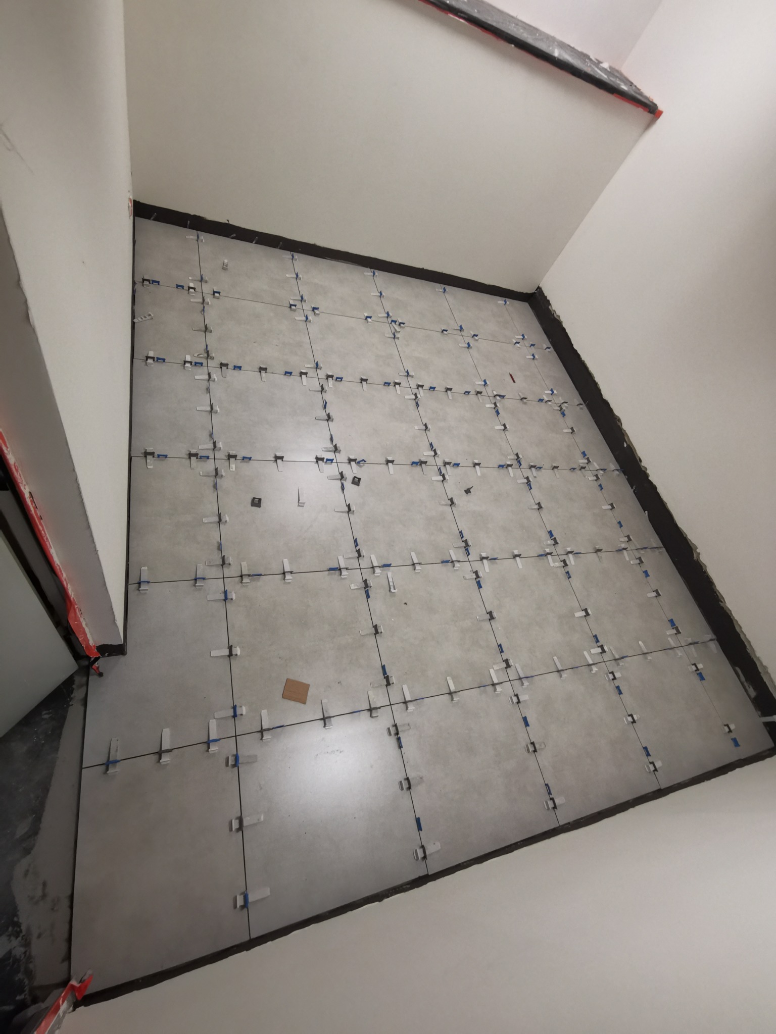 Szare, kwadratowe płytki podłogowe ułożone w regularną siatkę, z widocznymi krzyżykami dystansowymi w kolorze białym z niebieskimi elementami, na obrzeżach czarna fuga, ściany pomalowane na biało.