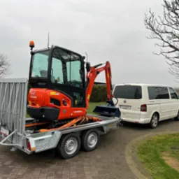 Pomarańczowa minikoparka Kubota KX019-4, zabezpieczona pasami transportowymi, na niskopodwoziowej przyczepie, zaparkowana za białym vanem Volkswagen Transporter na brukowanym podjeździe.