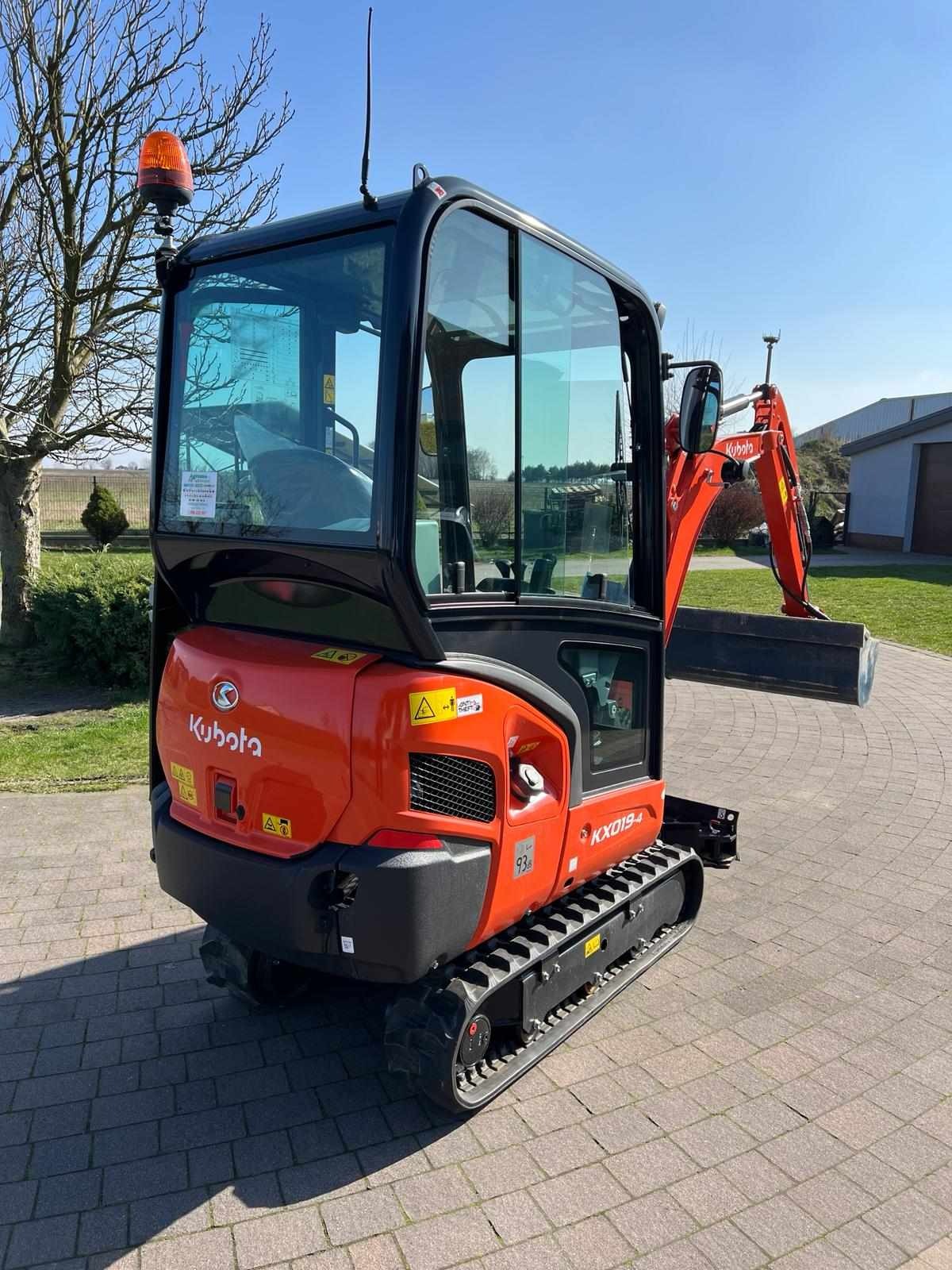 Pomarańczowa minikoparka Kubota KX019-4 na gąsienicach, z widoczną kabiną, lampą ostrzegawczą i łyżką, zaparkowana na kostce brukowej w słoneczny dzień.