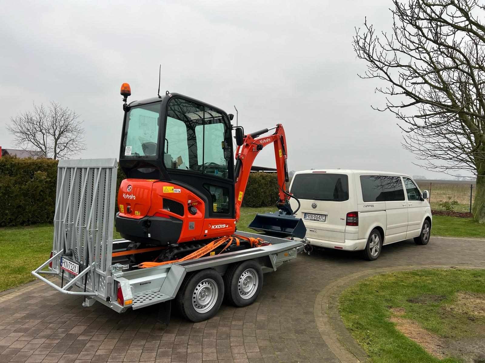 Pomarańczowa minikoparka Kubota KX019-4, zabezpieczona pasami transportowymi, na niskopodwoziowej przyczepie, zaparkowana za białym vanem Volkswagen Transporter na brukowanym podjeździe.