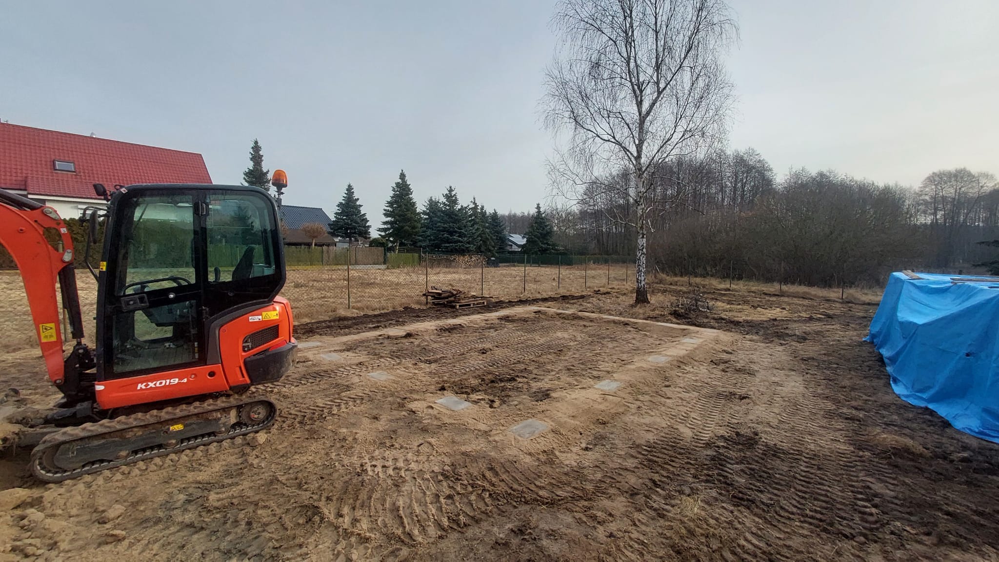Pomarańczowa minikoparka Kubota KX019-4 na placu budowy z wyznaczonym fundamentem, w tle drzewa i dom z czerwoną dachówką. Widoczne ślady opon na piasku.