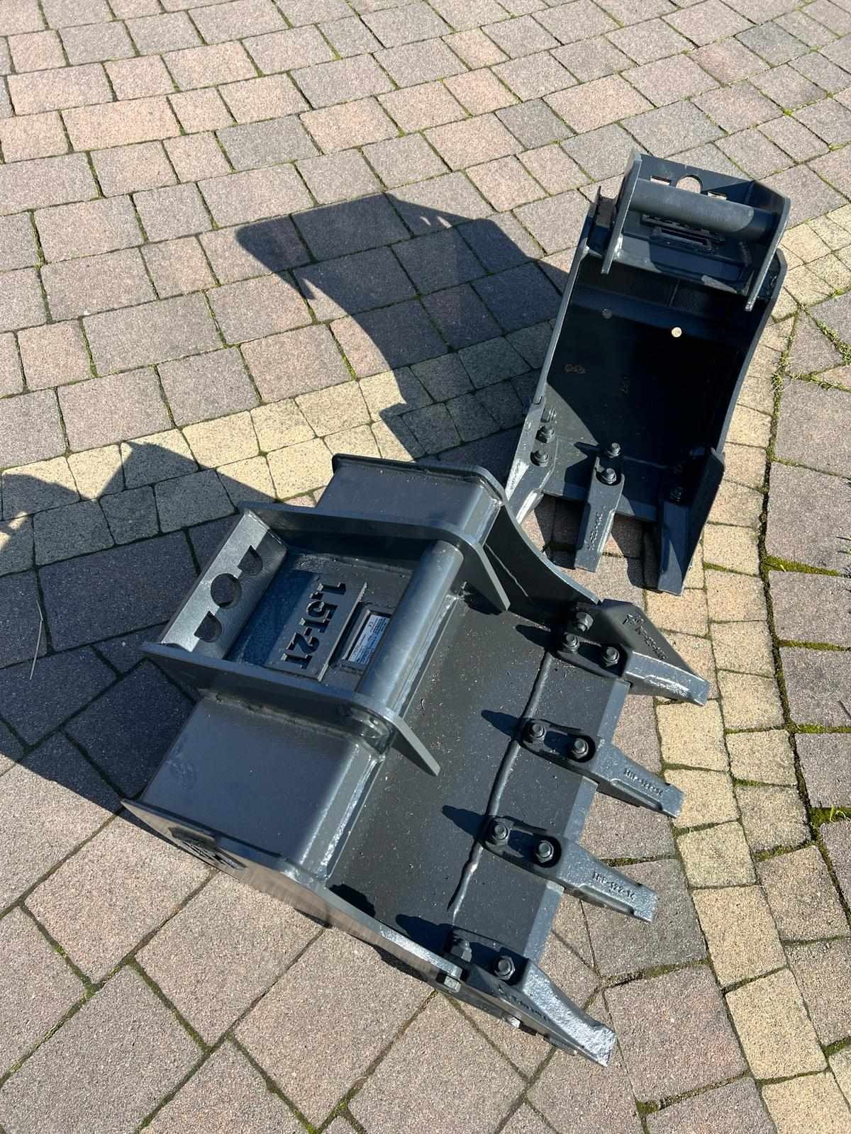 Dwa szare, metalowe łyżki do koparki leżące na kostce brukowej w słoneczny dzień. Na jednej z nich widoczny napis '1.5T-2T'.