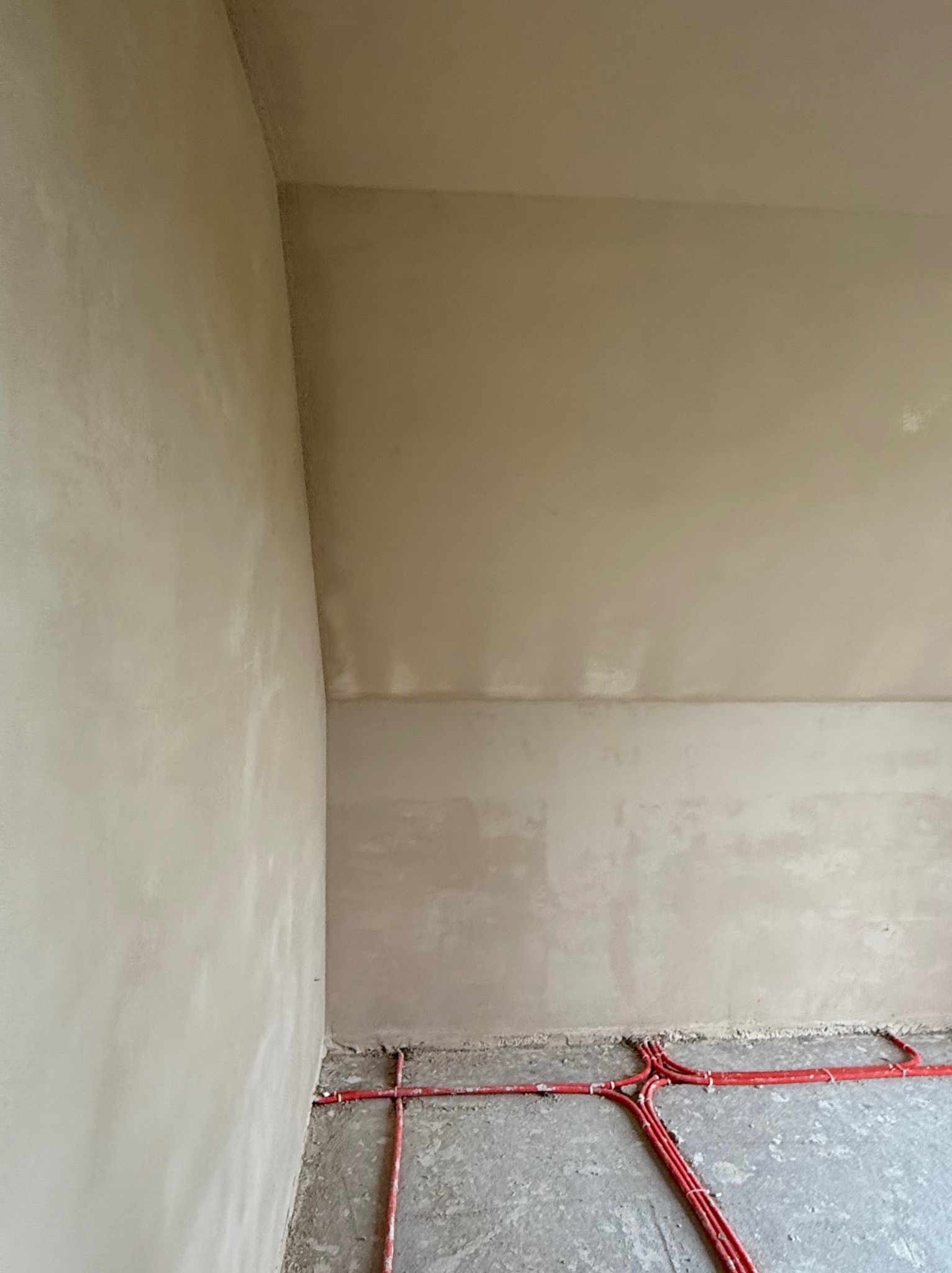 Świeżo położony tynk maszynowy na ścianach wewnętrznych, z widoczną instalacją ogrzewania podłogowego w kolorze czerwonym przy podłodze. Gładka powierzchnia tynku.