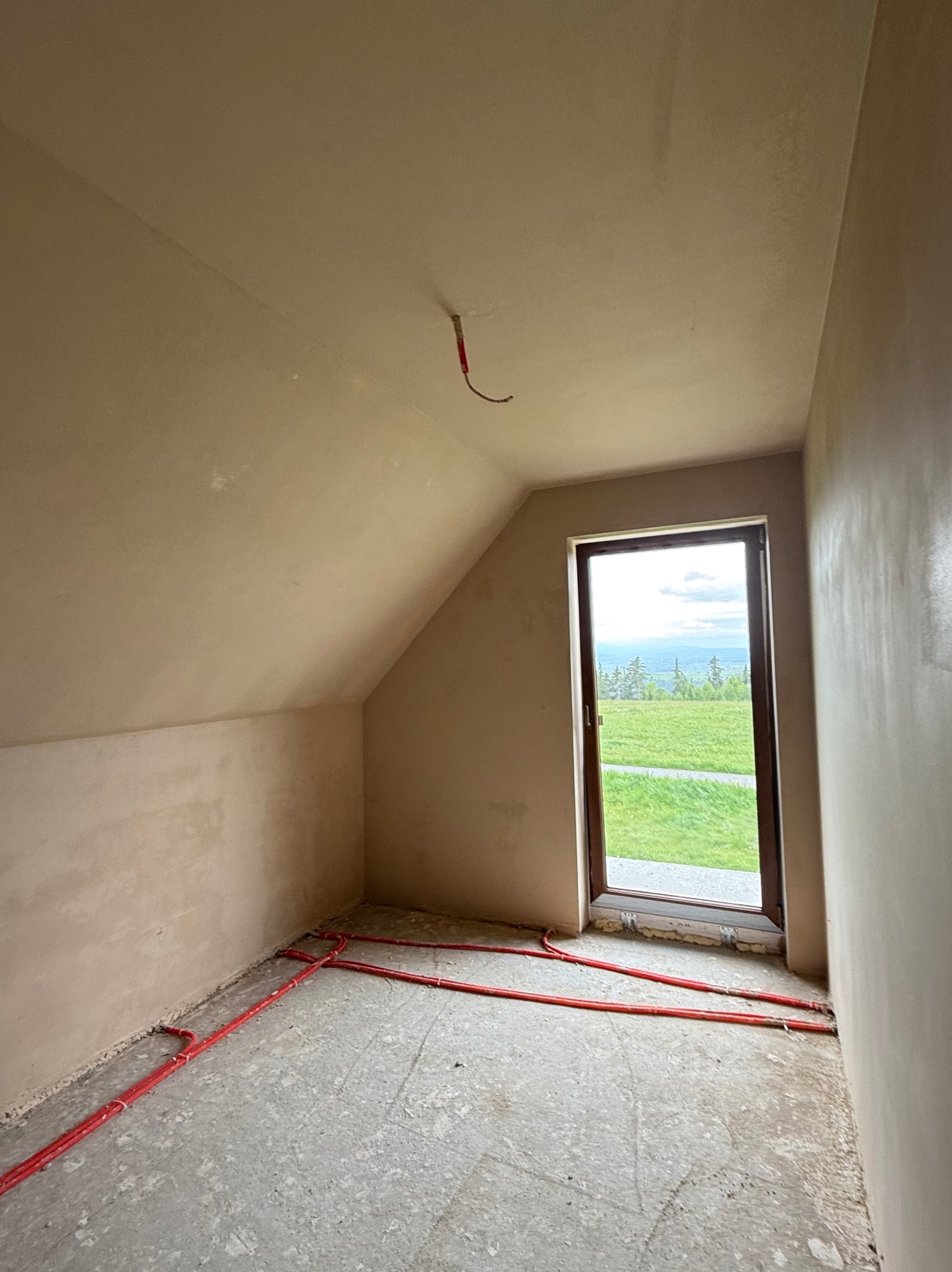Poddasze po tynkowaniu maszynowym, widoczne rury ogrzewania podłogowego przy ścianach i okno z widokiem na zieleń. Ściany gładkie, przygotowane do dalszych prac wykończeniowych.