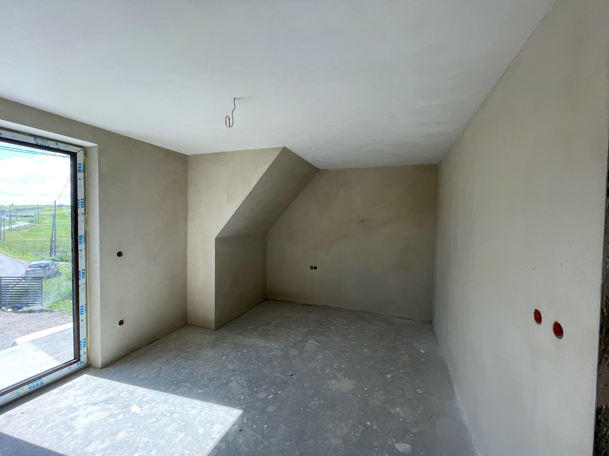 Wnętrze poddasza po tynkowaniu: gładkie, jasne ściany, surowa podłoga, okno z widokiem na zewnątrz. Widoczne puszki elektryczne w ścianach.