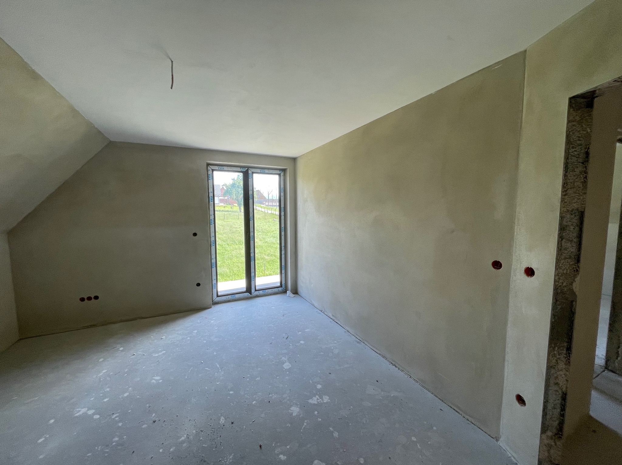 Pokój na poddaszu po tynkowaniu maszynowym: gładkie, jasne ściany, okno z widokiem na zieleń, surowa podłoga. Widoczne puszki elektryczne.