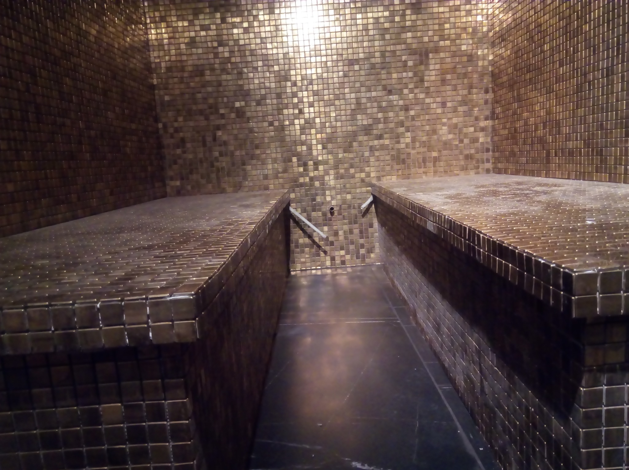 Wnętrze sauny wykończone drobną, złotą mozaiką, prezentujące dwa poziome siedziska z odpływem wody, oświetlone górnym światłem.