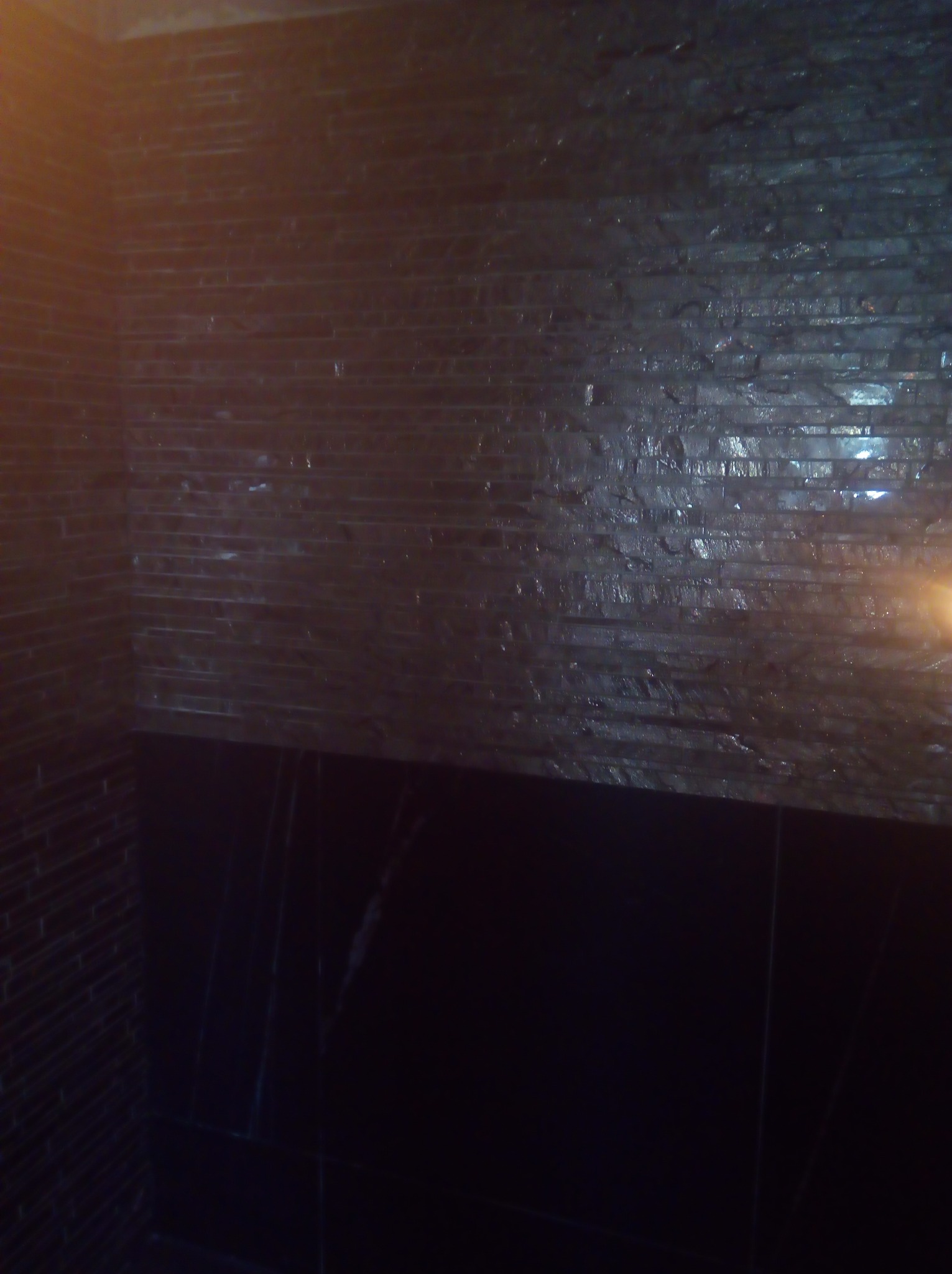 Ciemna ściana wyłożona błyszczącymi, wąskimi płytkami imitującymi kamień, z widocznymi poziomymi spoinami, poniżej fragment ciemnej, gładkiej powierzchni.