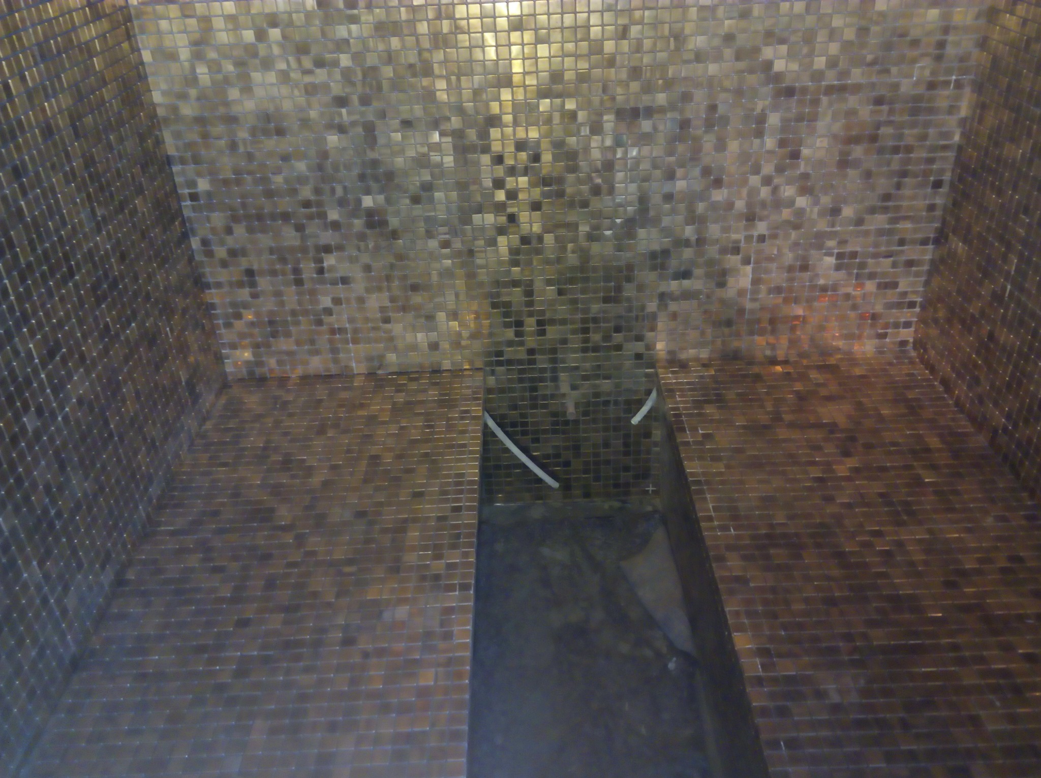 Wnętrze prysznica wyłożone mozaiką w odcieniach brązu i złota, z widocznymi białymi rurkami instalacji wodnej w górnej części.