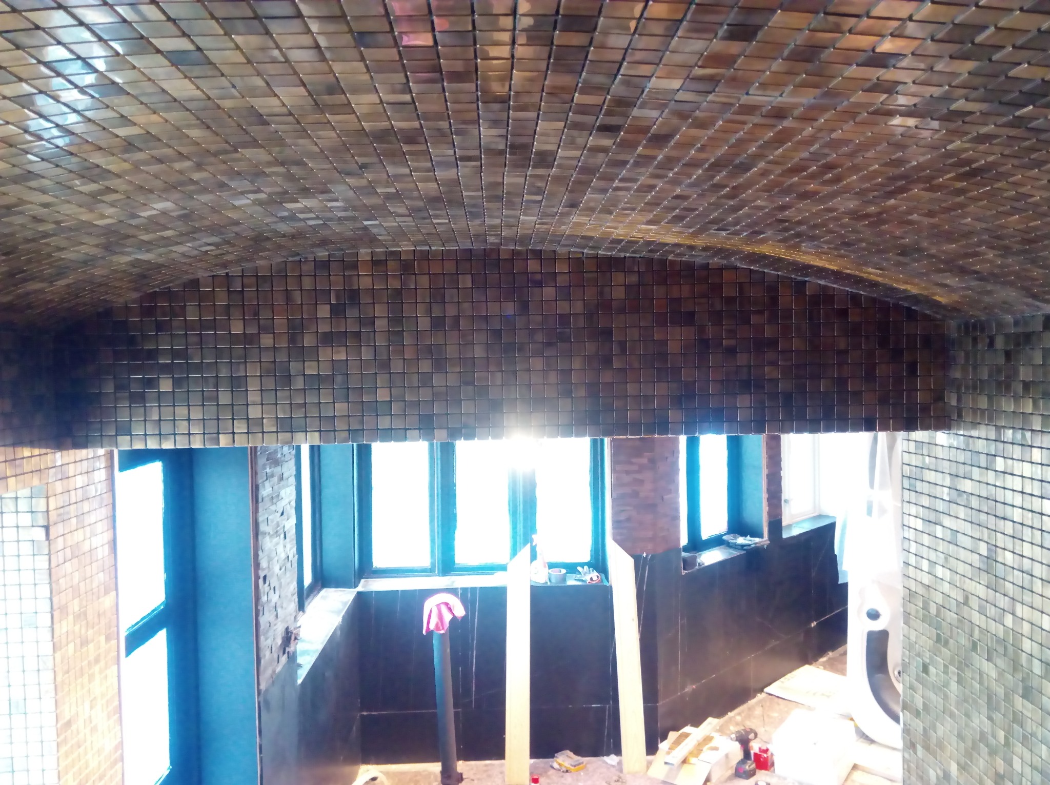 Mozaika z małych, metalicznych płytek pokrywająca sufit i ściany łazienki w trakcie remontu, widoczne okno i narzędzia pracy.