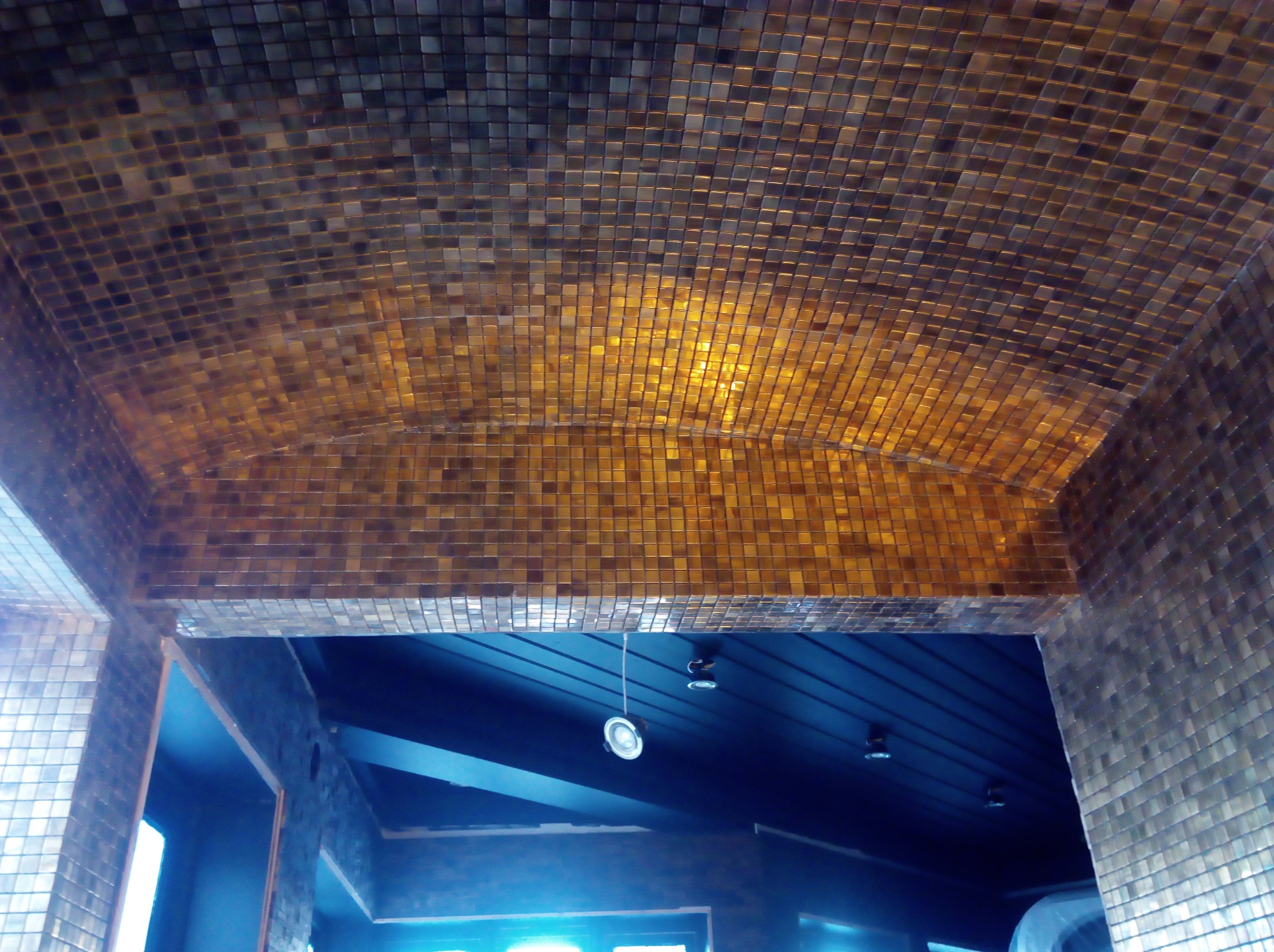 Sufit i ściany wykończone mozaiką z małych, błyszczących płytek w odcieniach złota i brązu, oświetlone punktowymi lampami, widok z dołu.