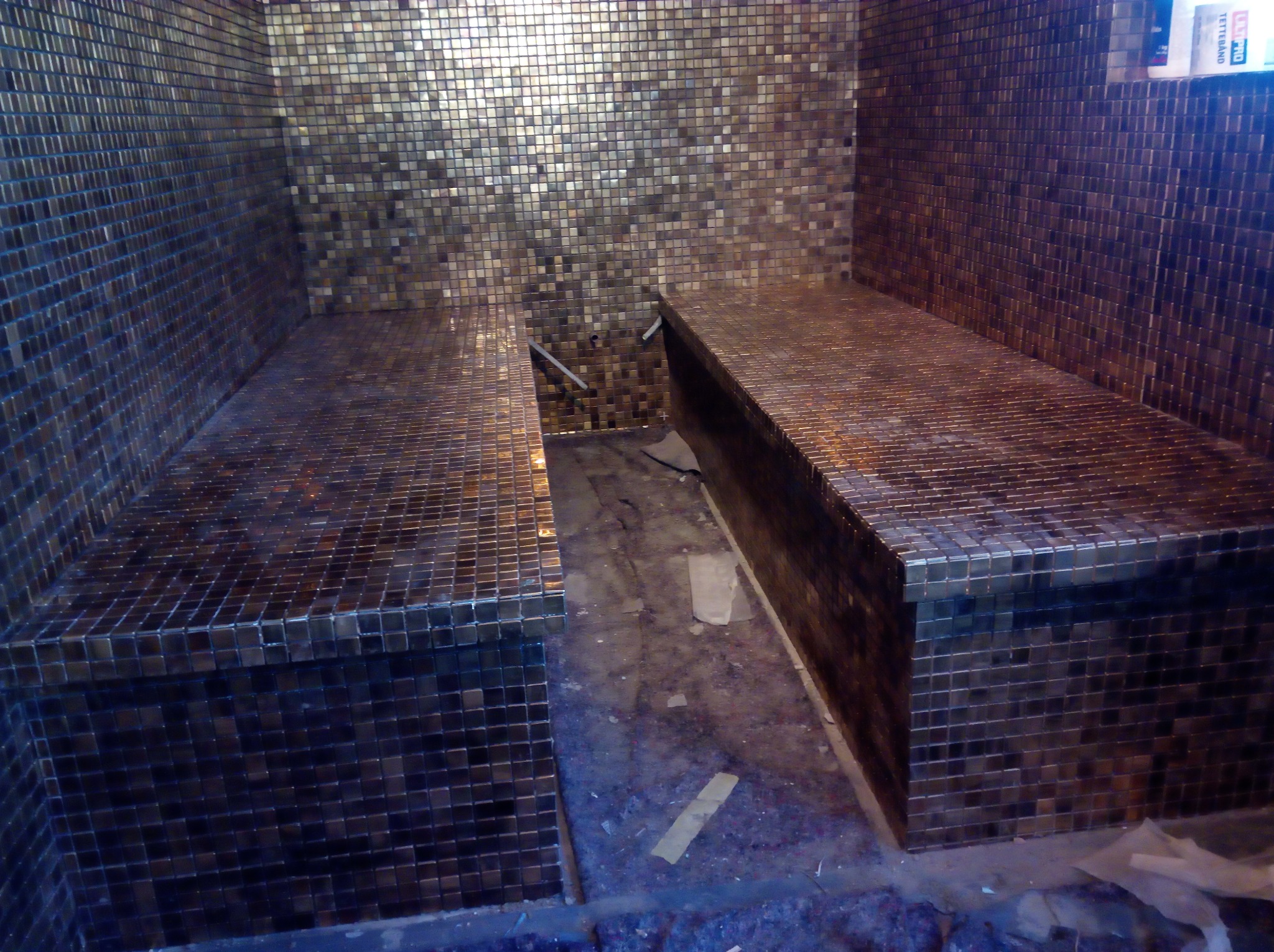 Dwa wyłożone mozaiką siedziska w parnej łaźni, ściany również pokryte mozaiką w odcieniach brązu i złota, widoczne niedociągnięcia na podłodze sugerujące etap prac.