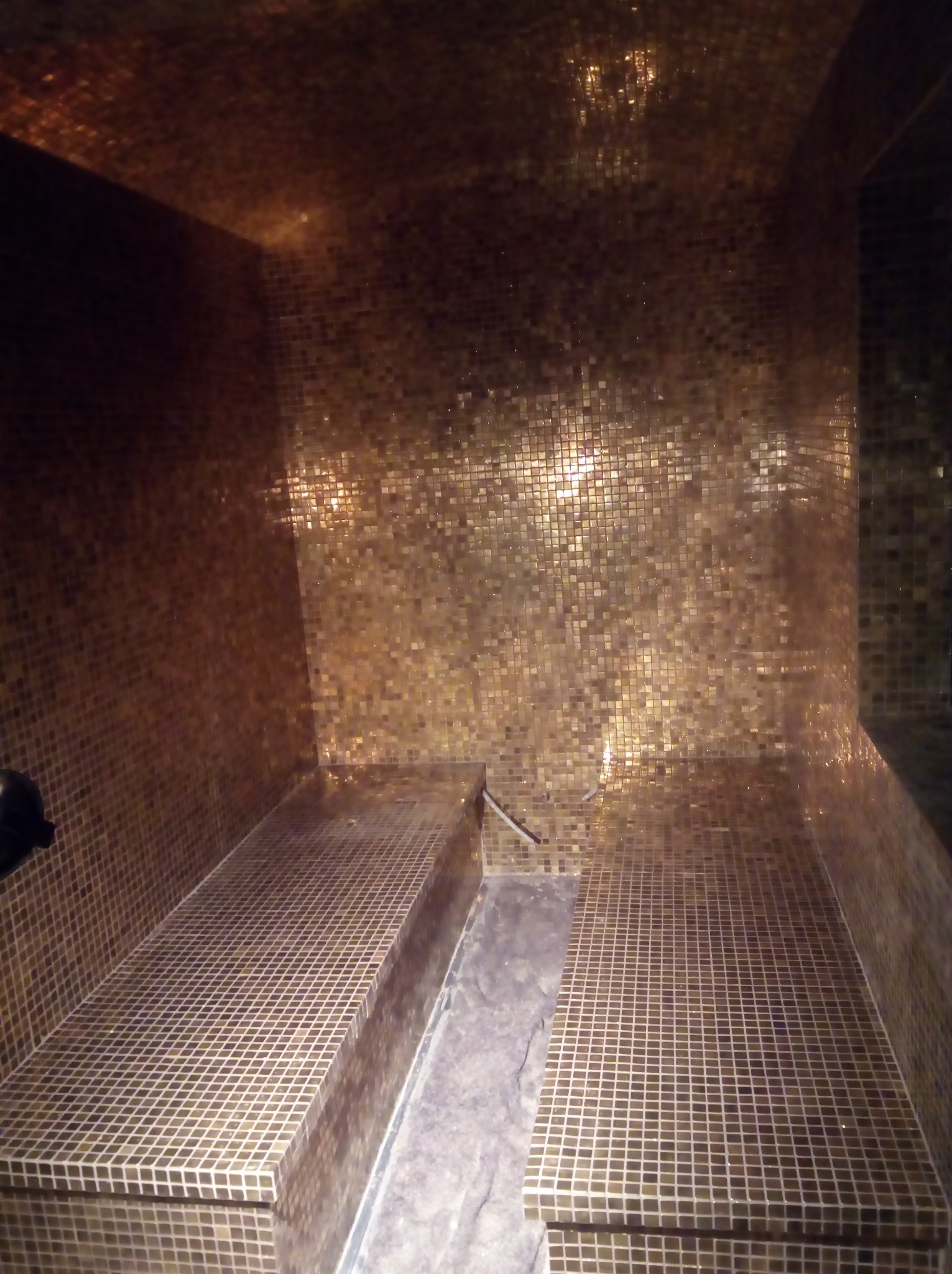 Wnętrze sauny wykończone złotą mozaiką, z dwoma poziomymi siedziskami również pokrytymi mozaiką, oświetlenie punktowe tworzy refleksy na ścianach.