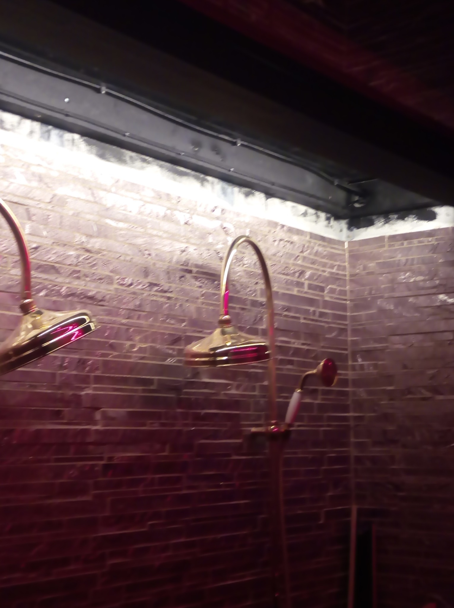 Dwa złote prysznice z czerwonymi akcentami zamontowane na ścianie z cegły w łazience oświetlonej czerwonym światłem.