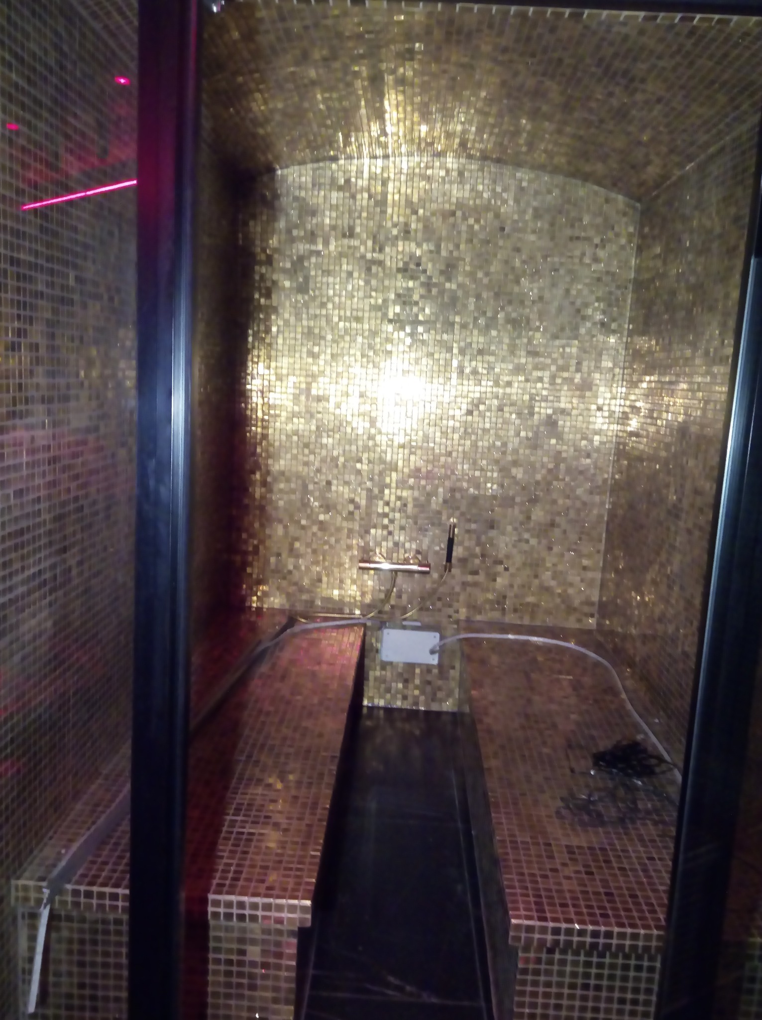 Wnętrze kabiny prysznicowej wykończonej złotą mozaiką, z dwoma siedziskami również pokrytymi mozaiką, widoczne czarne elementy wykończeniowe i oświetlenie.