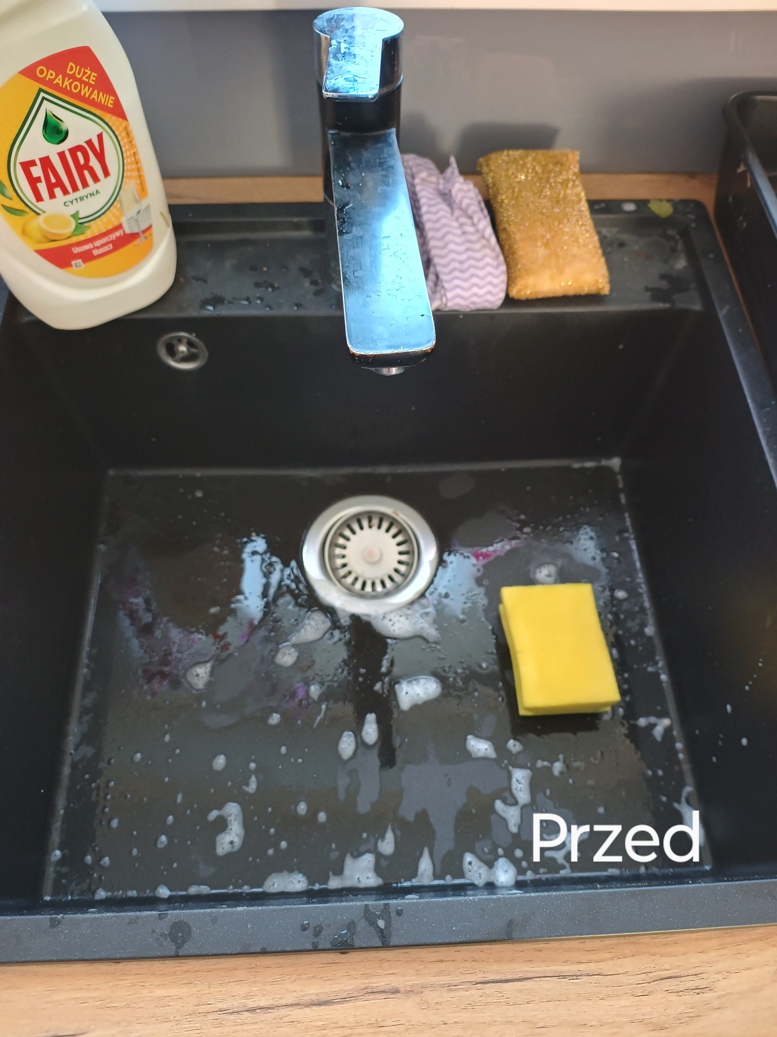 Czarny, zabrudzony zlew kuchenny z resztkami jedzenia i płynem do naczyń Fairy, gąbka do mycia naczyń oraz ściereczka w tle, widok z góry.
