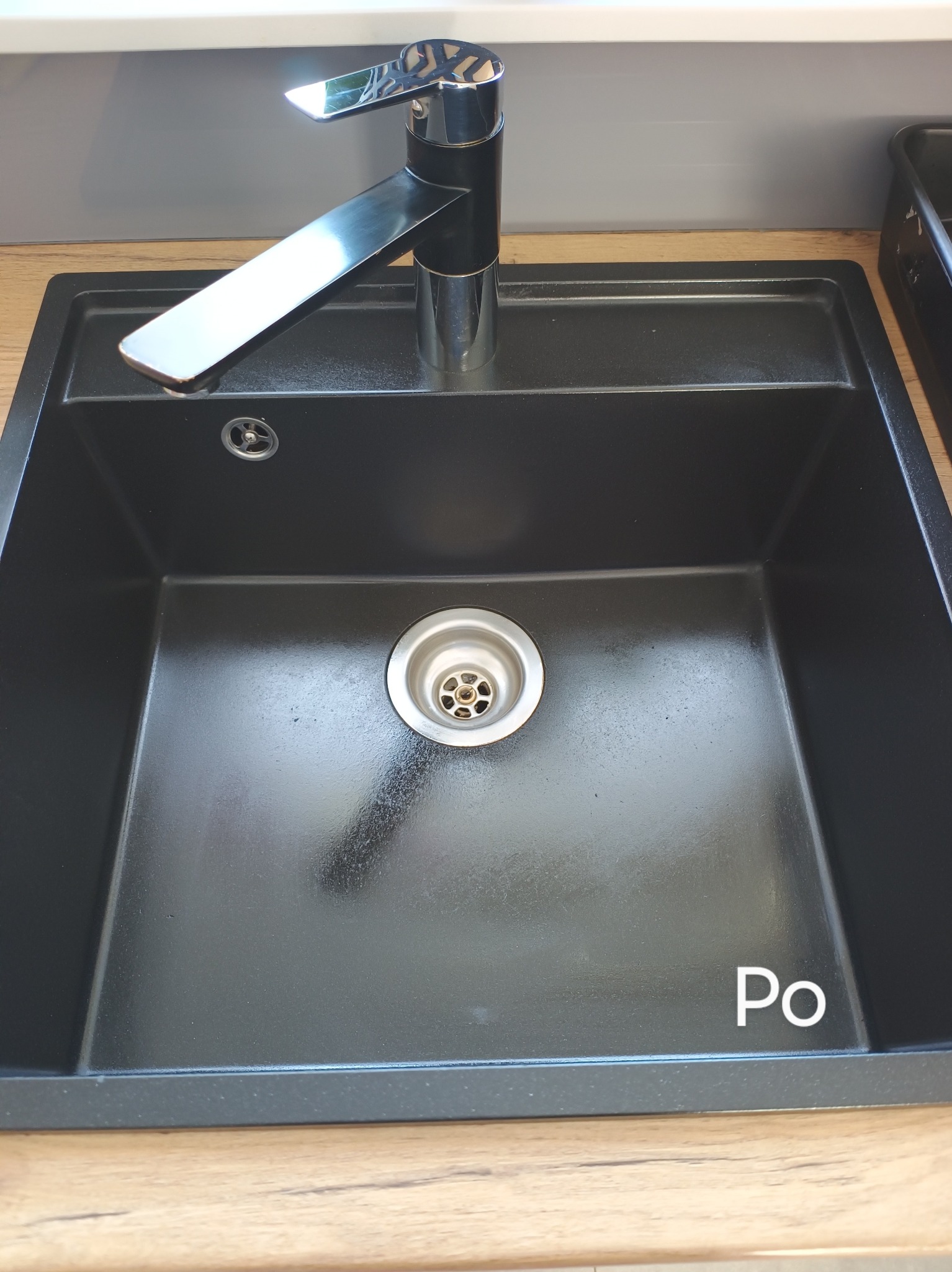 Czarny, kwadratowy zlew granitowy z chromowaną baterią, widoczny po czyszczeniu, z napisem 'Po' w prawym dolnym rogu.