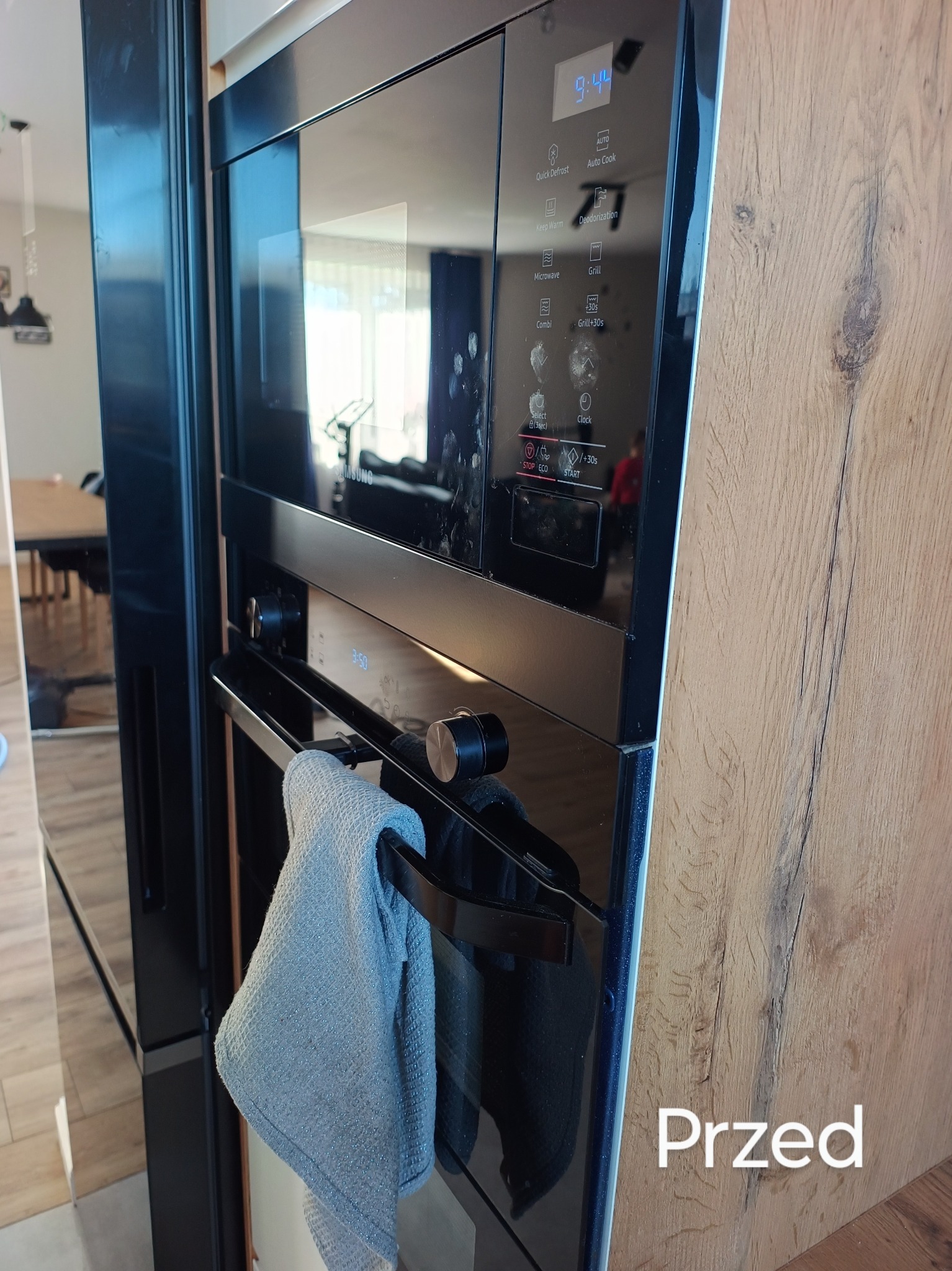 Czarny piekarnik z mikrofalówką, widoczne ślady użytkowania na szklanym froncie, na uchwycie wisi szara ściereczka, napis 'Przed' sugeruje stan przed czyszczeniem.