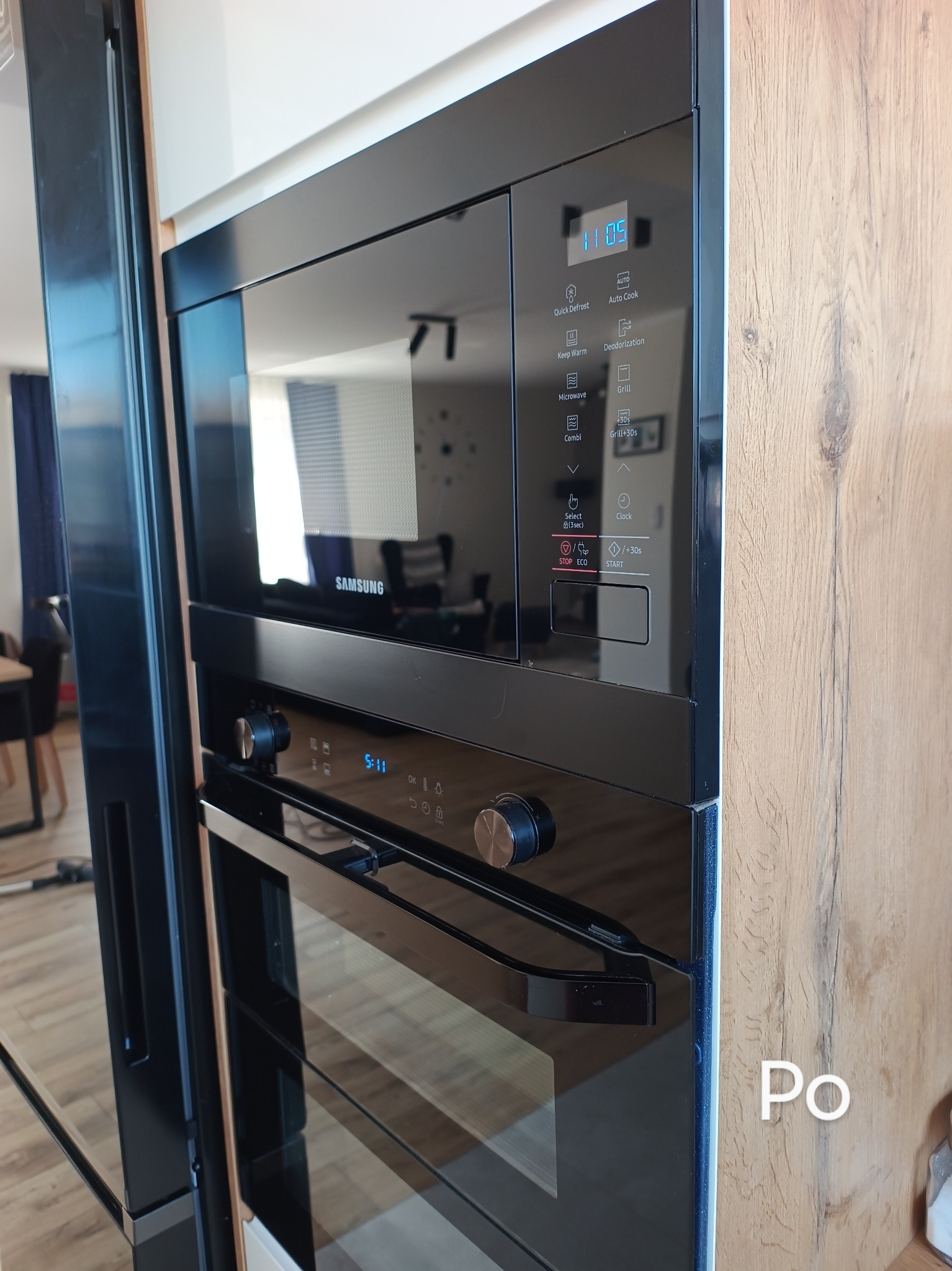 Wbudowany, czarny piekarnik i mikrofalówka Samsung z wyświetlaczem wskazującym godzinę 11:05, zamontowane w drewnianej zabudowie kuchennej, widok z boku.