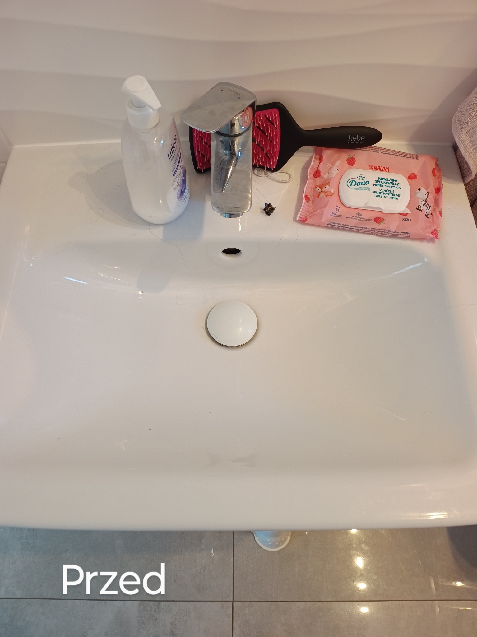 Biała umywalka z widocznymi śladami użytkowania i włosami, obok dozownik z mydłem, szczotka do włosów i opakowanie nawilżanych chusteczek Dada Malina przed czyszczeniem.