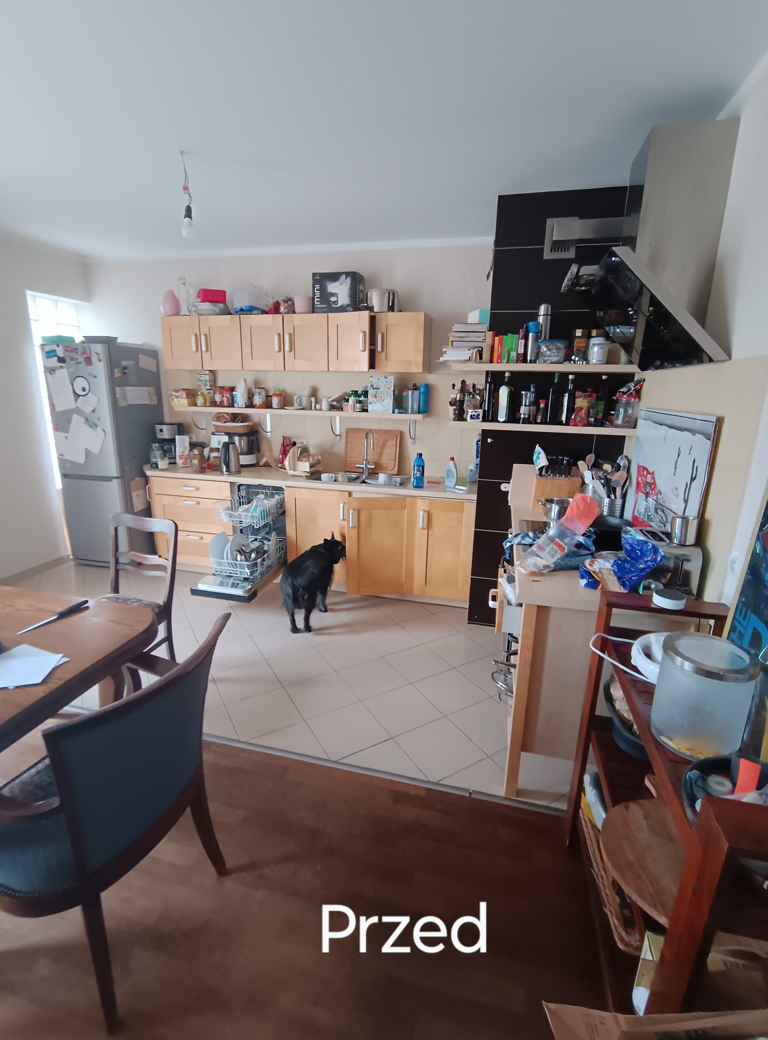 Zdjęcie przedstawia kuchnię w stanie 'przed', z widocznym bałaganem na blatach i otwartą zmywarką. Na pierwszym planie stół z krzesłami i czarny pies.