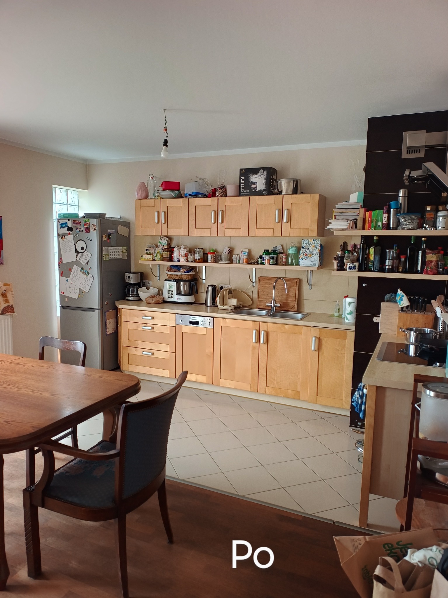 Jasna kuchnia po sprzątaniu: drewniane szafki, czyste blaty, lśniąca podłoga z płytek. Widok na stół i krzesła. Napis 'Po' sugeruje efekt końcowy usługi.