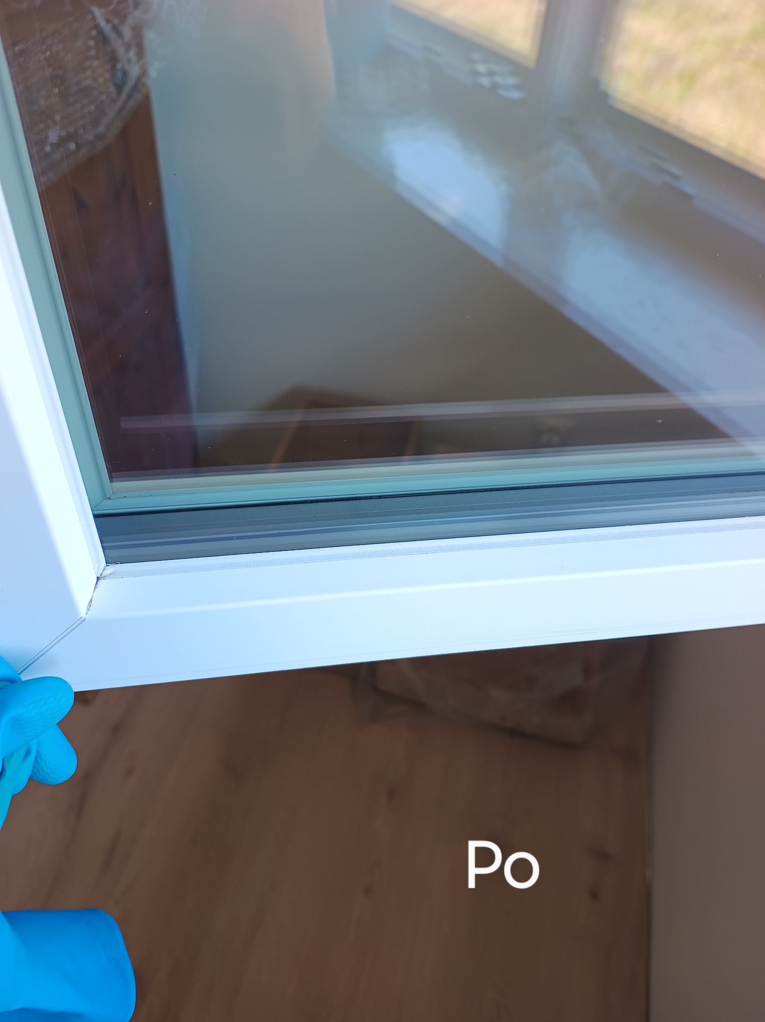 Czyste okno PCV po umyciu, widok z bliska z fragmentem dłoni w niebieskiej rękawicy. Na dole napis 'Po'.