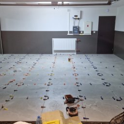 MatMax - Szare płytki podłogowe w trakcie układania, z użyciem klipsów i klinów poziomujących, widoczne narzędzia glazurnicze i butelka wody, ściany pomalowane dwukolorowo.