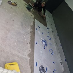 MatMax - Układanie jasnych płytek na ścianie; widoczny glazurnik w trakcie pracy, poziomica, przyssawka do płytek oraz system poziomowania płytek.