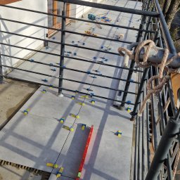 MatMax - Kładzenie szarych płytek na balkonie z użyciem poziomicy i niebiesko-żółtych krzyżyków dystansowych, widok z góry na częściowo wykończoną powierzchnię.