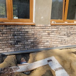 MatMax - Montaż elewacji z cegły klinkierowej na ścianie budynku, widoczne okna z drewnianą ramą, podłoże w trakcie prac ziemnych z widoczną folią kubełkową i fragmentami cegieł.