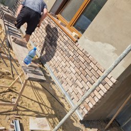 MatMax - Pracownik na rusztowaniu układa cegły klinkierowe na elewacji budynku, widoczna butelka wody i narzędzia na desce rusztowania.