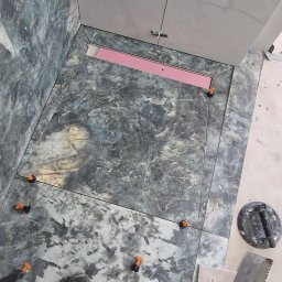 MatMax - Montaż płytek marmurkowych w łazience, widoczny odpływ liniowy z różową membraną, przyssawka do płytek i narzędzia glazurnicze, zastosowane klipsy poziomujące.