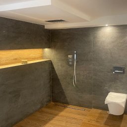 MatMax - Nowoczesna łazienka z ciemnymi, matowymi płytkami imitującymi kamień na ścianach, drewnianą podłogą i wnęką z drewnianą półką. Uwagę zwraca minimalistyczna bateria prysznicowa i toaleta...