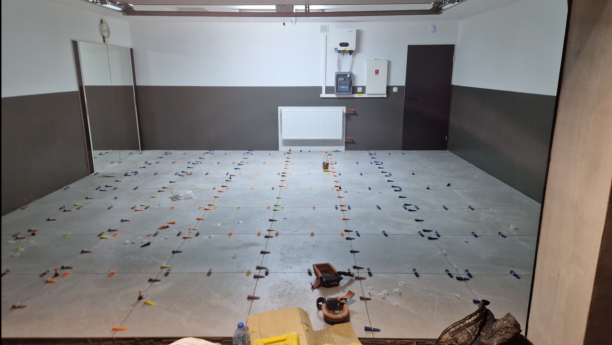 Szare płytki podłogowe w trakcie układania, z użyciem klipsów i klinów poziomujących, widoczne narzędzia glazurnicze i butelka wody, ściany pomalowane dwukolorowo.