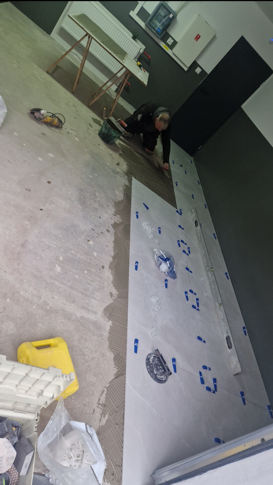 Układanie jasnych płytek na ścianie; widoczny glazurnik w trakcie pracy, poziomica, przyssawka do płytek oraz system poziomowania płytek.