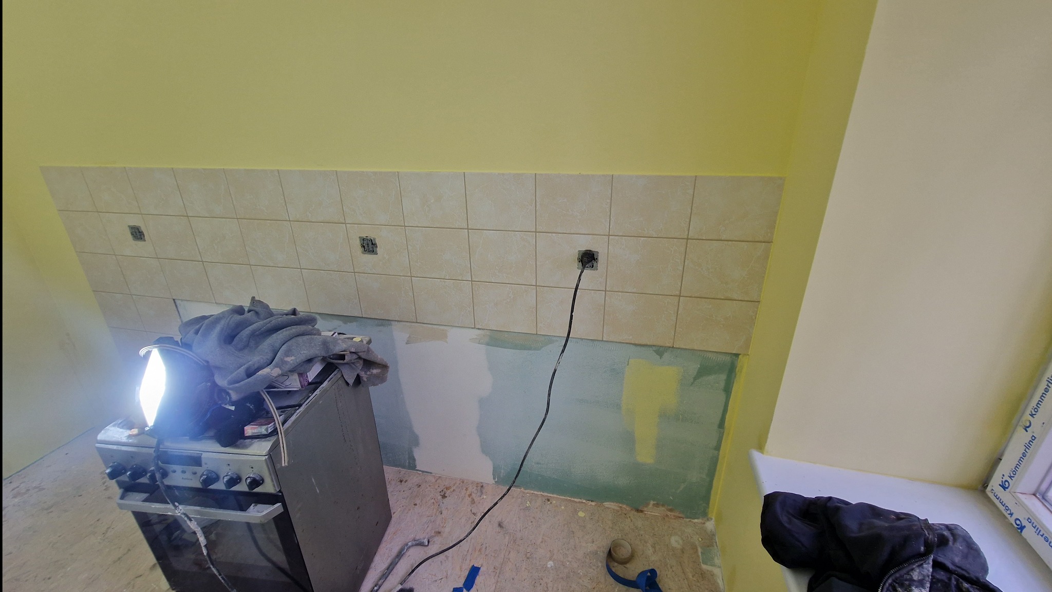 Niedokończony remont kuchni: częściowo położone beżowe płytki nad blatem, odsłonięte puszki elektryczne, prowizoryczne oświetlenie na kuchence, ślady farby na ścianach i podłodze.