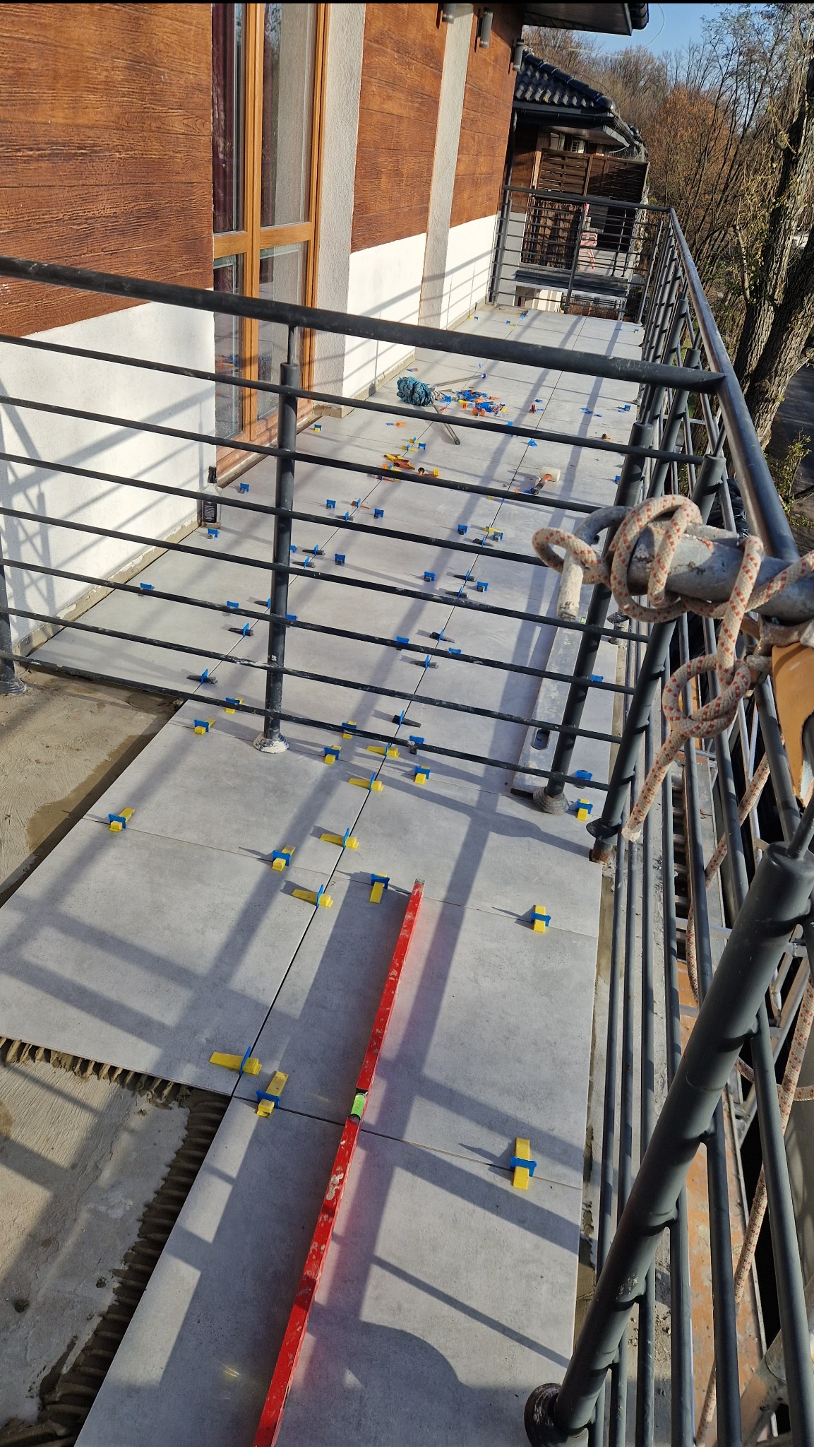 Kładzenie szarych płytek na balkonie z użyciem poziomicy i niebiesko-żółtych krzyżyków dystansowych, widok z góry na częściowo wykończoną powierzchnię.