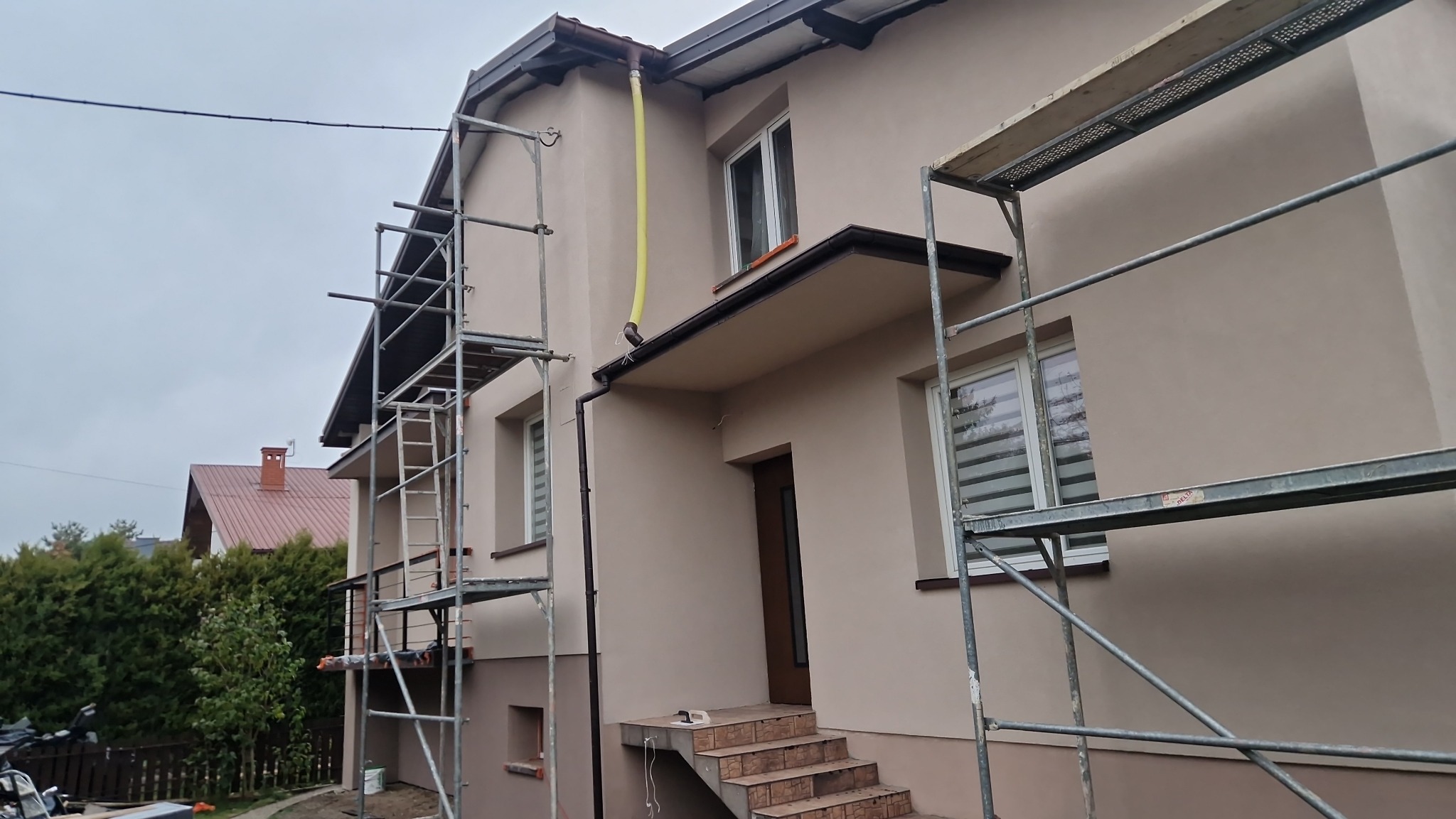 Elewacja domu w trakcie prac wykończeniowych, widoczne rusztowania i żółty wąż odprowadzający wodę z rynny, świeżo nałożona gładź w odcieniach beżu.