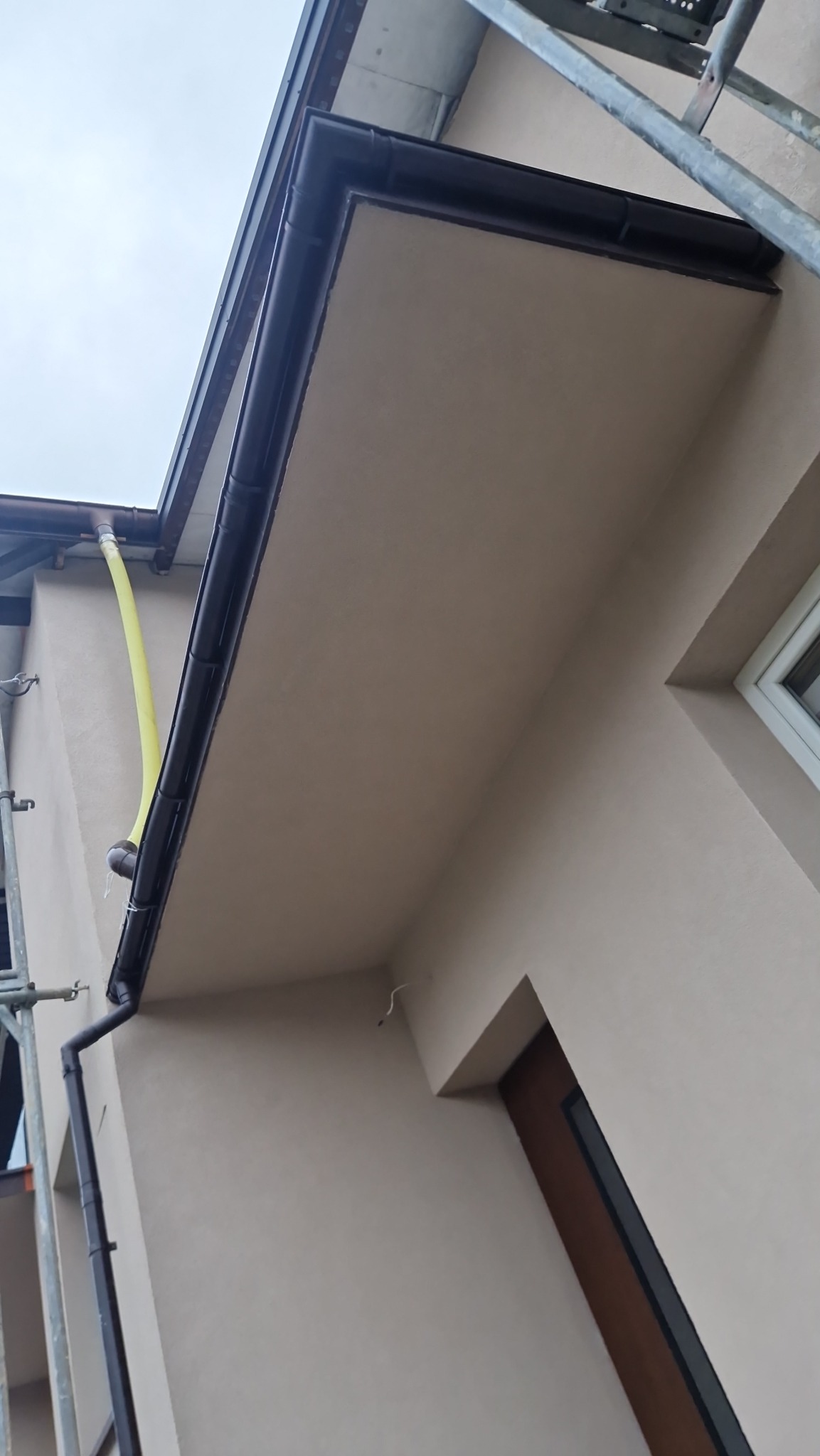 Beżowa elewacja budynku z ciemnymi rynnami, widoczne rusztowanie i żółty wąż odprowadzający wodę, gładka powierzchnia ściany.