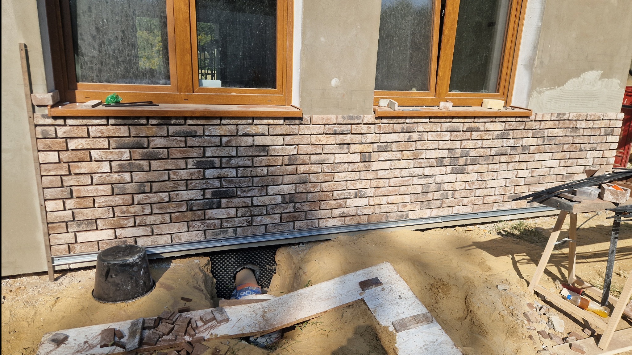 Montaż elewacji z cegły klinkierowej na ścianie budynku, widoczne okna z drewnianą ramą, podłoże w trakcie prac ziemnych z widoczną folią kubełkową i fragmentami cegieł.