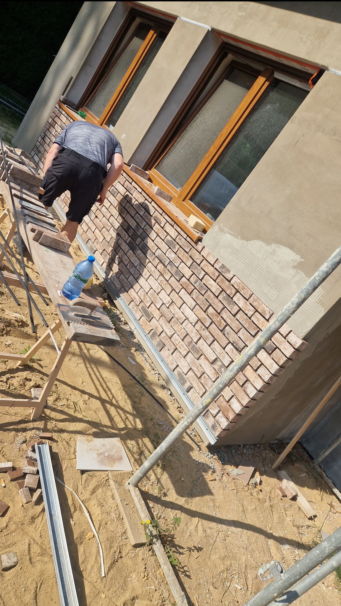 Pracownik na rusztowaniu układa cegły klinkierowe na elewacji budynku, widoczna butelka wody i narzędzia na desce rusztowania.