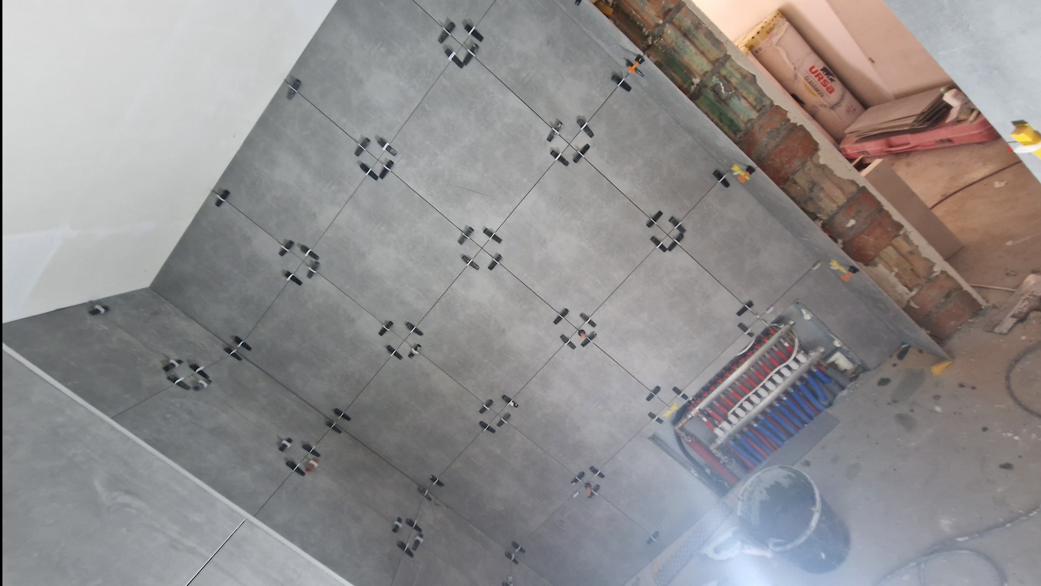 Szare płytki podłogowe układane w kwadraty z użyciem klipsów poziomujących, widoczne rury ogrzewania podłogowego, surowy mur i worki z zaprawą w tle.