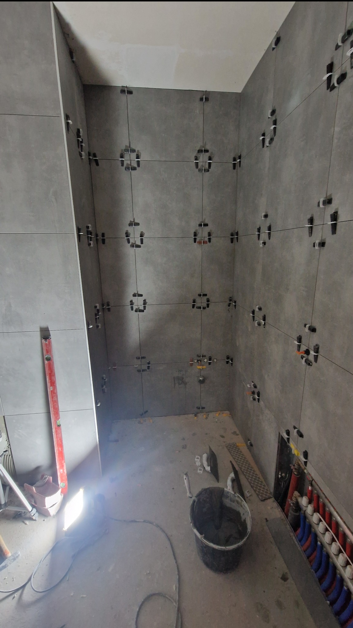 Szare płytki łazienkowe w trakcie montażu, z użyciem systemu poziomowania płytek, widoczny poziomica i wiadro z zaprawą.