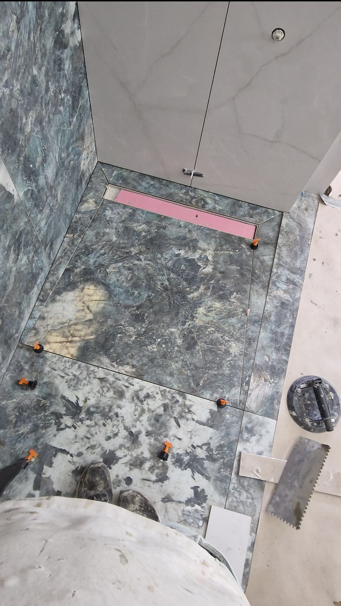 Montaż płytek marmurkowych w łazience, widoczny odpływ liniowy z różową membraną, przyssawka do płytek i narzędzia glazurnicze, zastosowane klipsy poziomujące.