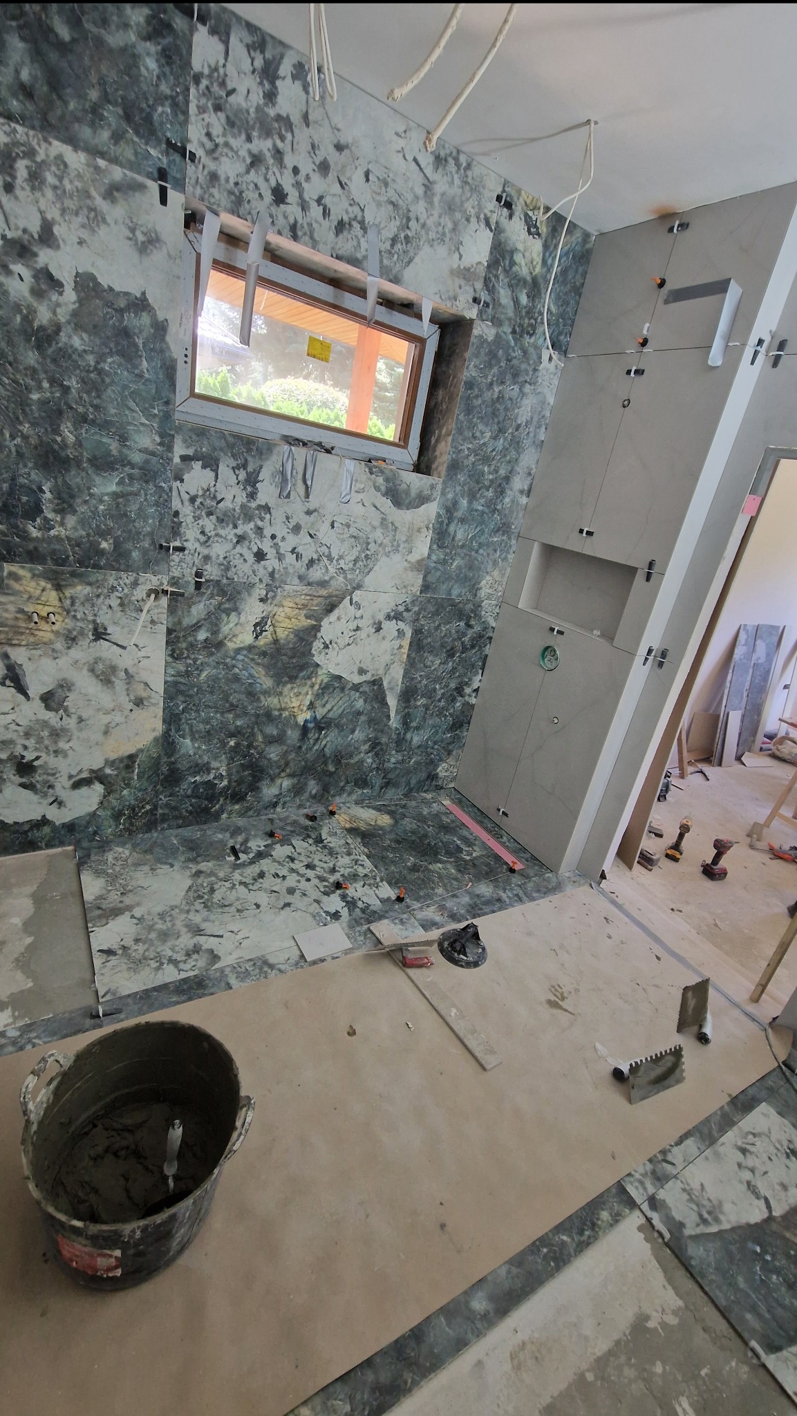 Wnętrze łazienki w trakcie prac wykończeniowych, widoczna ściana z płytek imitujących marmur, obok fragment ściany z szarych płytek, na podłodze wiadro z zaprawą i narzędzia glazurnicze.