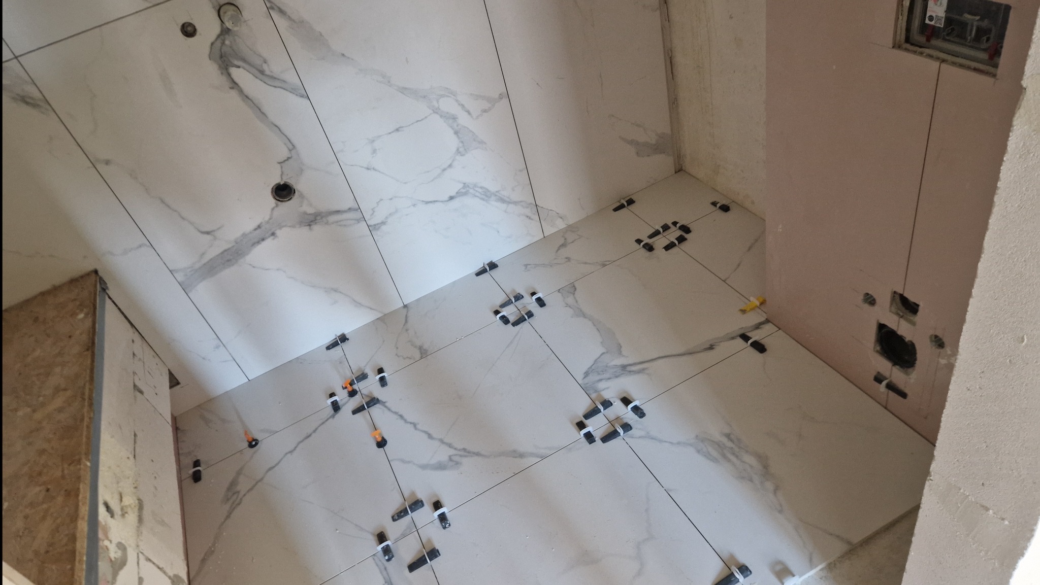 Układanie białych płytek imitujących marmur na podłodze i ścianach łazienki z użyciem systemów poziomowania płytek, widoczne otwory na instalacje elektryczne.