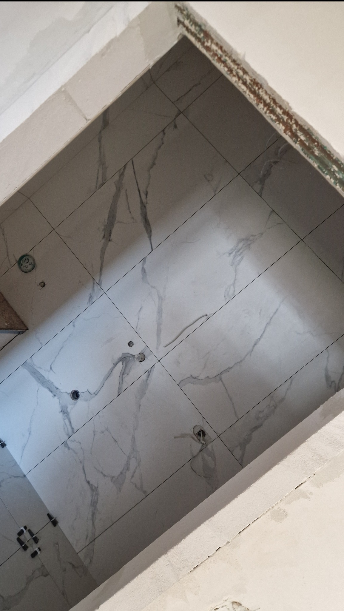 Wnętrze w trakcie prac: ułożone białe płytki imitujące marmur na ścianie, widoczne otwory na instalacje i niedokończone obramowanie otworu drzwiowego.